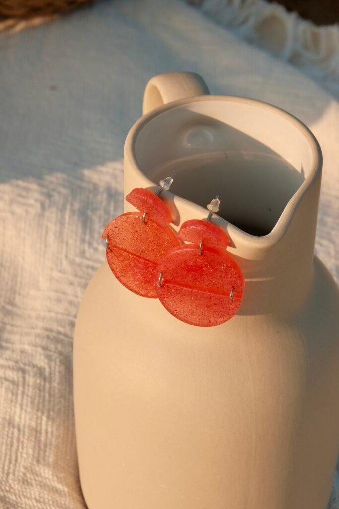 Hibiscus Amarillo red handcrafted earrings from the Costa Brava in Bloom collection, displayed on a clear glass water jug in natural light.